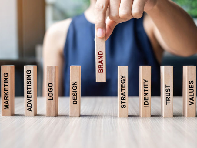 Brand-Equity-Blog Person holding a wooden block labelled "brand", placing it between other marketing-labelled blocks