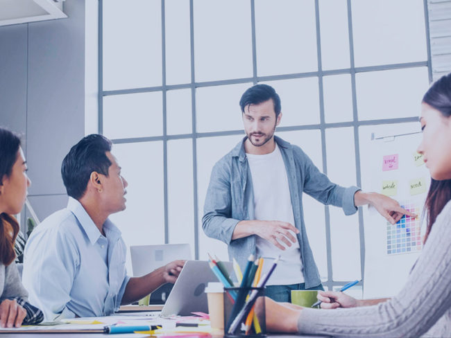 brand-strategist-skills-blog Meeting of four people, one stands and points to a whiteboard