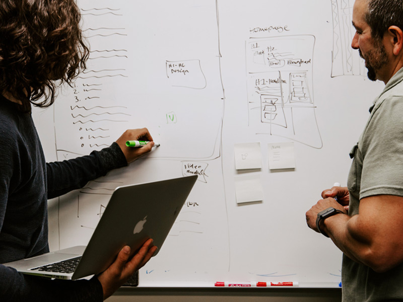 brand-strategist-tools-blog A person writes on a whiteboard as they hold a laptop as someone stands beside her