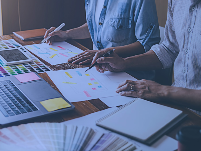 what-does-a-brand-strategist-do-blog Two people holding pens and sitting at a desk, surrounded by paper, notebooks, and a laptop