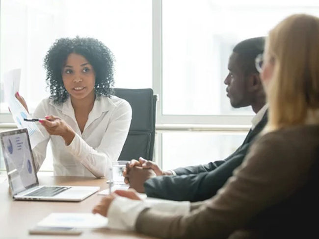 brand-strategist-benefits-blog Woman gesturing to a paper as she talks to two people in a business meeting