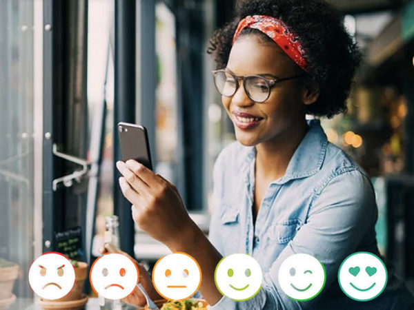 A woman holding her phone as she sits inside, a row of ranging emotions of emoji icons below her