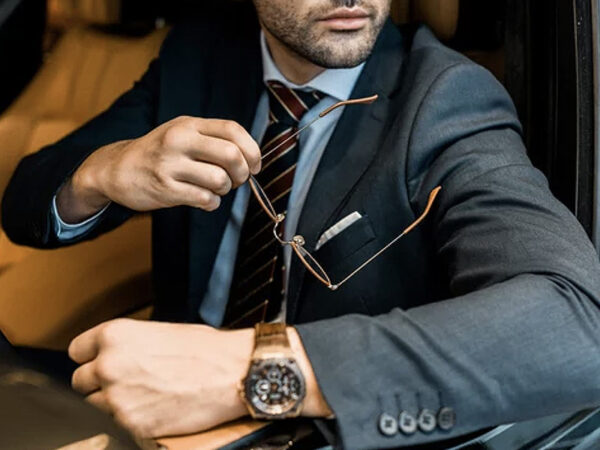 Man in a business suit sitting in a car, propping his elbow on the car door and holding glasses