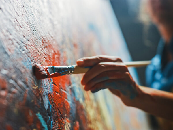 Hand holding a paintbrush against a canvas with multicolored paintstrokes